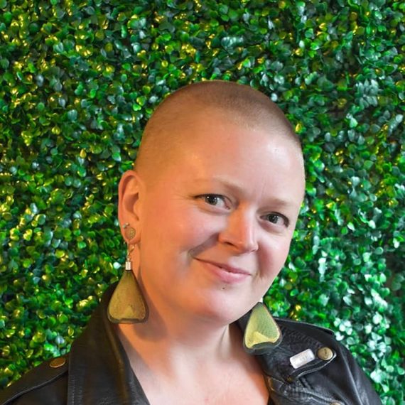 Sarah smiling wearing a black leather jacket and large triangle earrings.