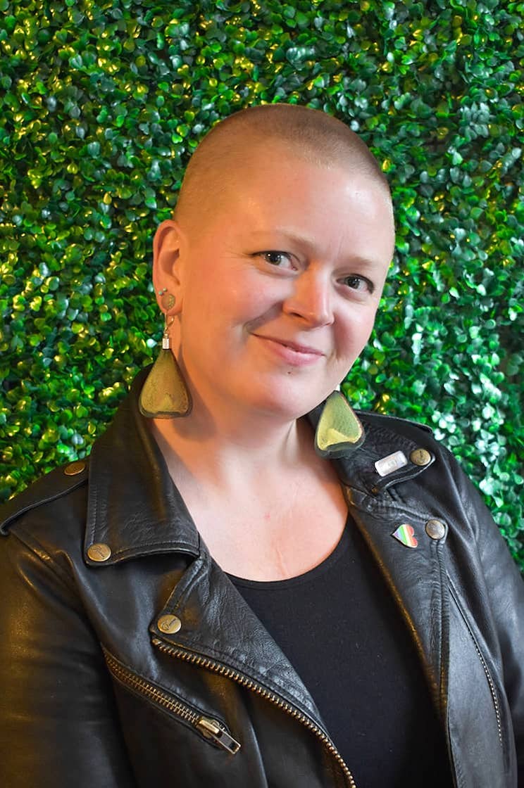 Sarah smiling wearing a black leather jacket and large triangle earrings.