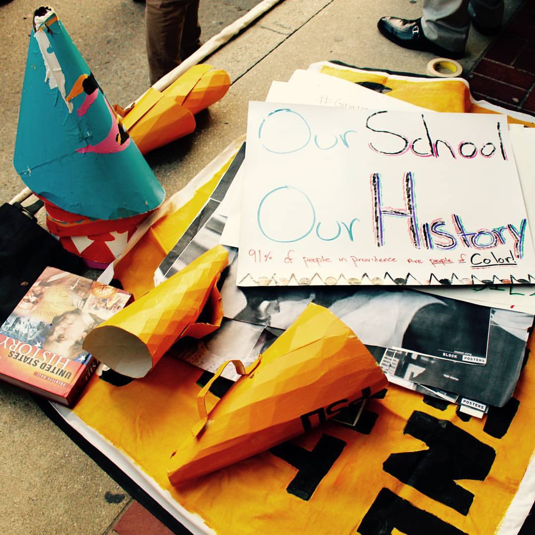 Assorted ephemera on the floor: a handmade poster that reads "Our School, Our History" and "91% of people in Providence are people of color!", a U.S. history book, homemade speaker cones, and other posters.